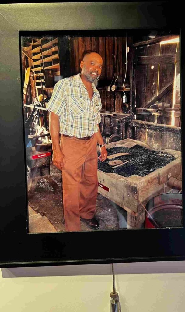 A framed photograph of a smiling Brandon Briggs standing next to the working coal forge inside the blacksmith shop.