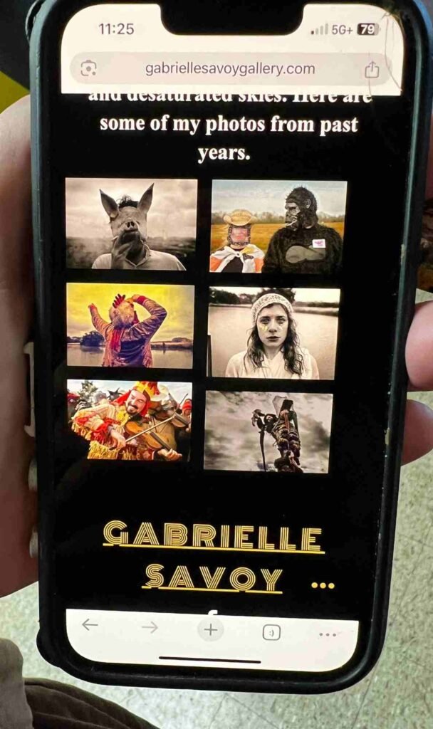 A hand holds a scratched iPhone displaying a website titled gabriellesavoygallery.com. The screen shows six artistic photographs arranged in a grid with the text “and desaturated skies. Here are some of my photos from past years” above them and the gold stylized name GABRIELLE SAVOY below. The phone shows the time 11:25 and 5G+ signal with 79% battery.