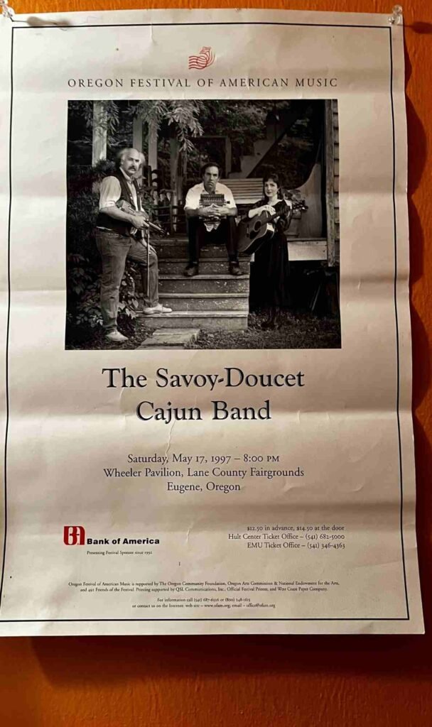 A vintage promotional poster for the Oregon Festival of American Music featuring a black‑and‑white photograph of three musicians from The Savoy‑Doucet Cajun Band—one with a guitar, one with an accordion, and one with another guitar—posing on outdoor steps