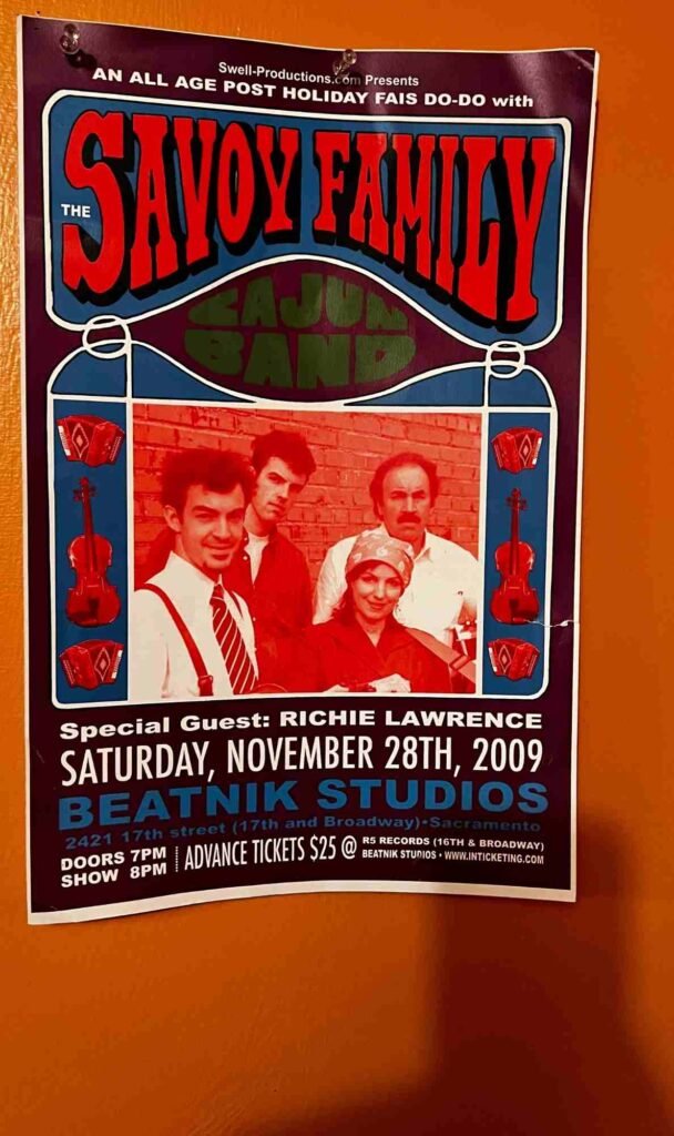 A vintage concert poster for an all‑age post‑holiday Fais Do‑Do event featuring the Savoy Family Cajun Band with special guest Richie Lawrence. The poster shows a red‑tinted photo of four band members framed by blue lanterns with violins and accordions. It advertises the show on Saturday, November 28th, 2009 at Beatnik Studios in Sacramento.