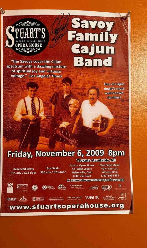 The photo shows a red concert poster for the Savoy Family Cajun Band at Stuart's Opera House in Nelsonville, Ohio, on Friday, November 6, 2009 at 8 pm. It features a photo of three band members with instruments and lists ticket prices and venue information. The poster includes sponsor logos and a website URL at the bottom.