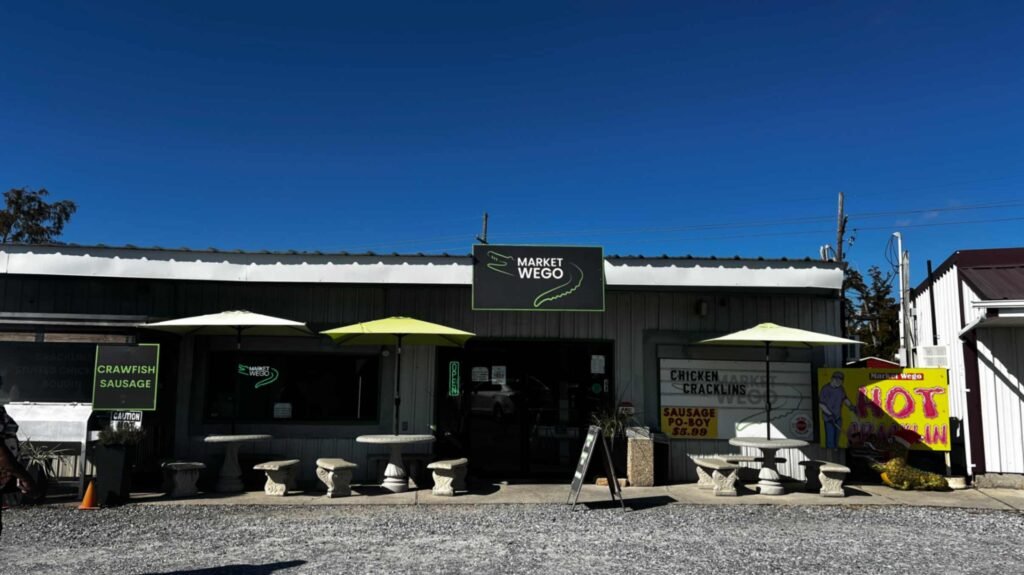 The exterior of "Market Wego," a white corrugated metal building with a dark sign featuring a green alligator logo. Three yellow umbrellas shade stone outdoor seating areas, and colorful signs advertise local favorites like crawfish sausage and chicken cracklings.