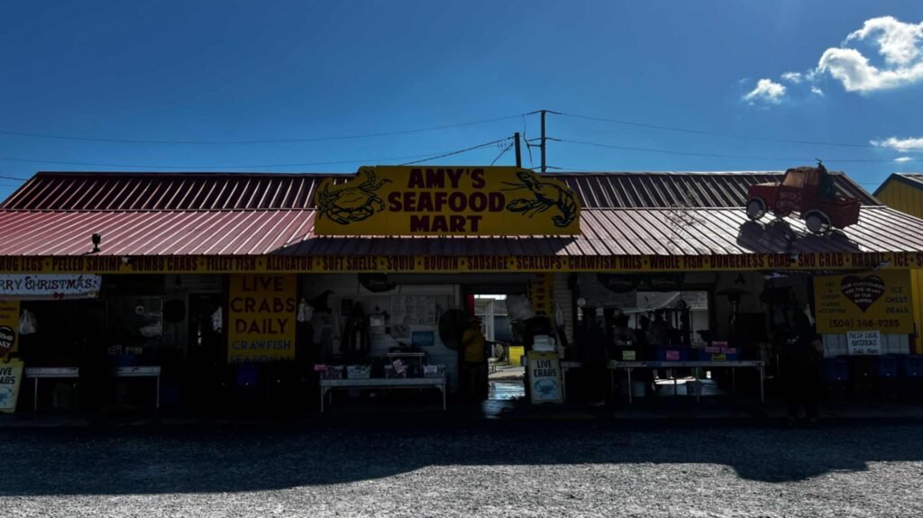 A wide shot of Amy’s Seafood Mart under a red corrugated metal roof. A large yellow sign with a crab and shrimp logo sits atop the building, while the eaves list various offerings like "Frog Legs," "Boudin," and "Scallops."