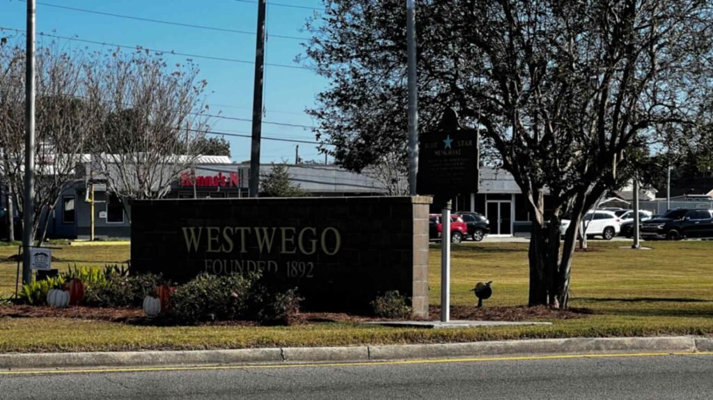 A dark stone monument sign on a manicured lawn reading "WESTWEGO FOUNDED 1892" in gold lettering. A Blue Star Memorial marker stands nearby under a leafy tree against a clear blue sky.