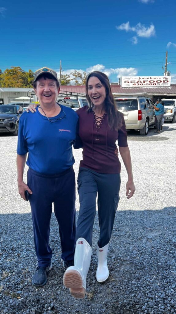 A smiling man in a blue shirt and camouflage hat stands next to a woman in a maroon top and white shrimp boots. They are standing on a gravel lot with cars and "Jonathan's Seafood" sign in the background.