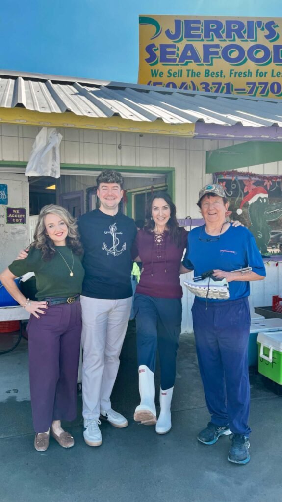 Four people standing together smiling in front of "Jerri’s Seafood". One woman wears white shrimp boots and a man in a blue shirt holds a grey sneaker. A Christmas-themed alligator decoration is visible in the background.