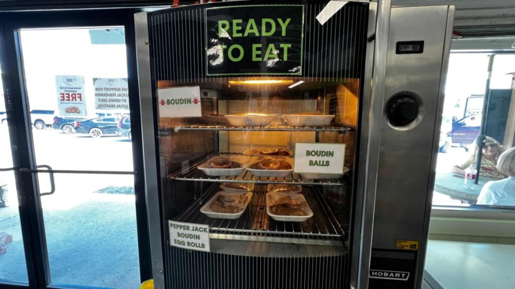 A heated food display case inside a market with a sign reading "READY TO EAT." Inside are trays of "Boudin Balls," "Pepper Jack Boudin Egg Rolls," and other local Cajun specialties.