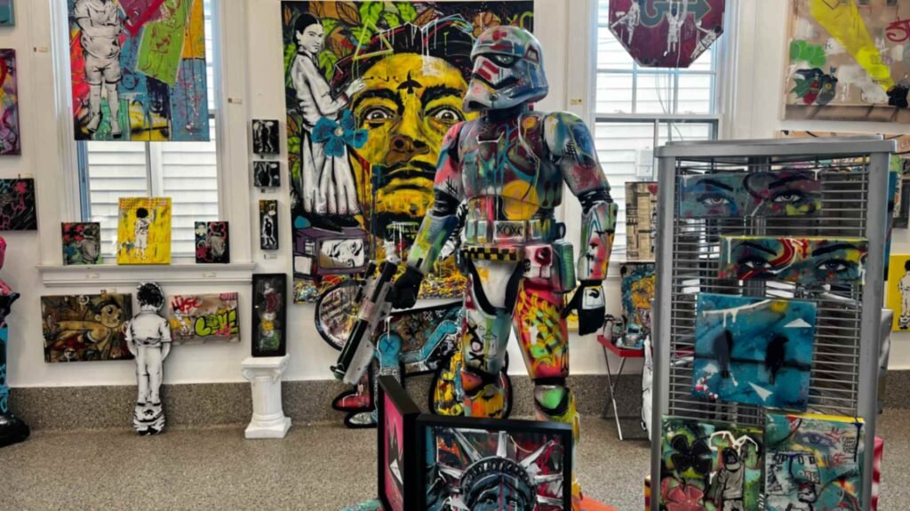 An interior view of the gallery featuring a life-sized Stormtrooper statue covered in colorful graffiti-style paint, surrounded by various pop art canvases and urban sculptures.