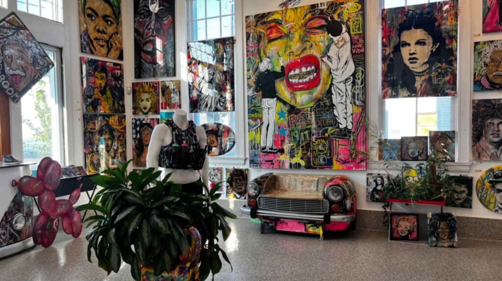 A spacious gallery room with high ceilings and large windows, filled with floor-to-ceiling artwork, a headless mannequin in a tactical vest, and a bench made from the front of a vintage car.