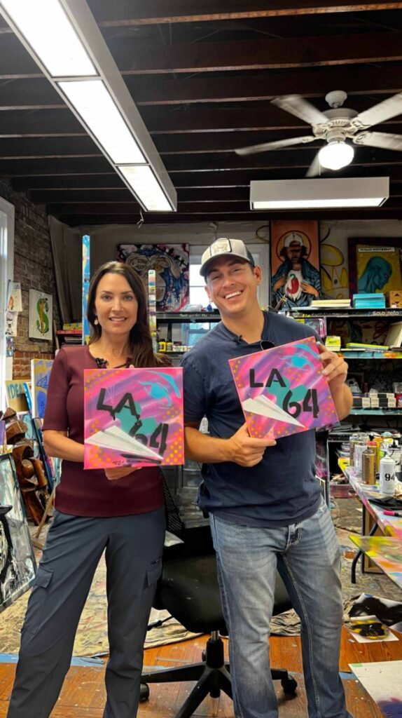 Joshua Wingerter and a woman standing in his art studio, both smiling and holding matching square canvases that feature "LA 64" text and a paper plane design over a pink and purple background.
