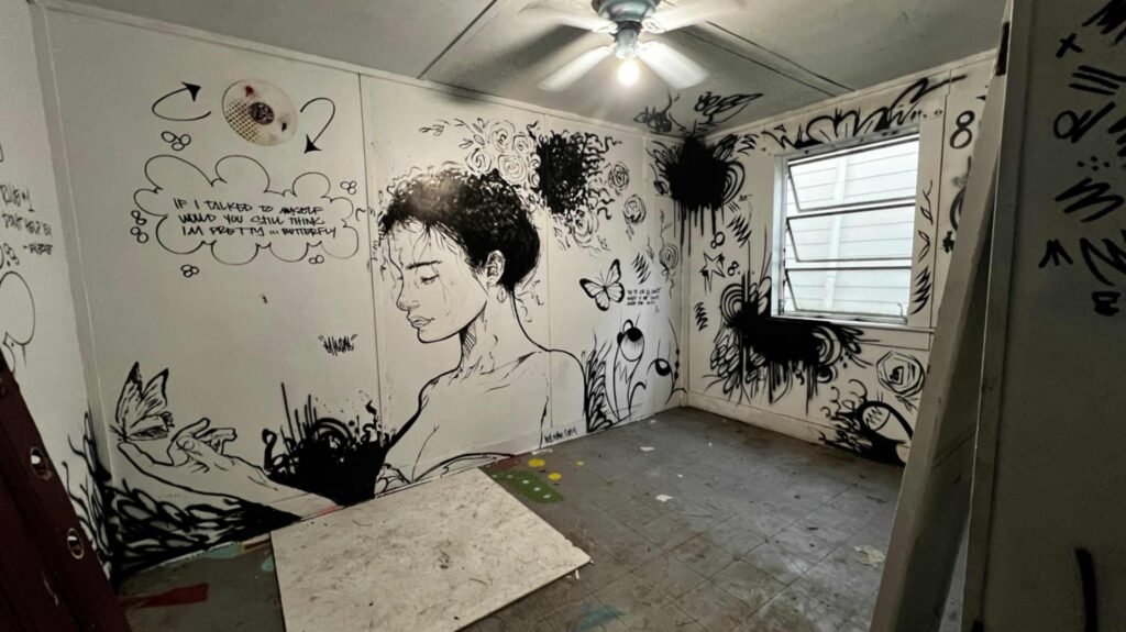 A minimalist room featuring a large black and white wall mural of a woman with flowers in her hair, surrounded by graffiti-style text and butterflies.