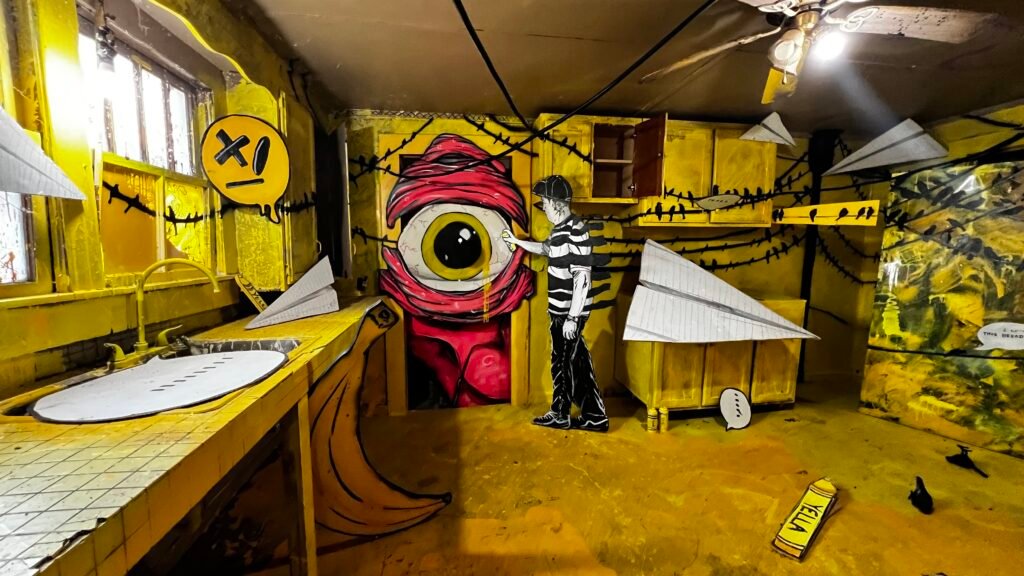 An immersive art installation of a kitchen entirely covered in yellow paint, featuring graffiti-style 2D cutouts of a giant eye, a banana, and paper planes.