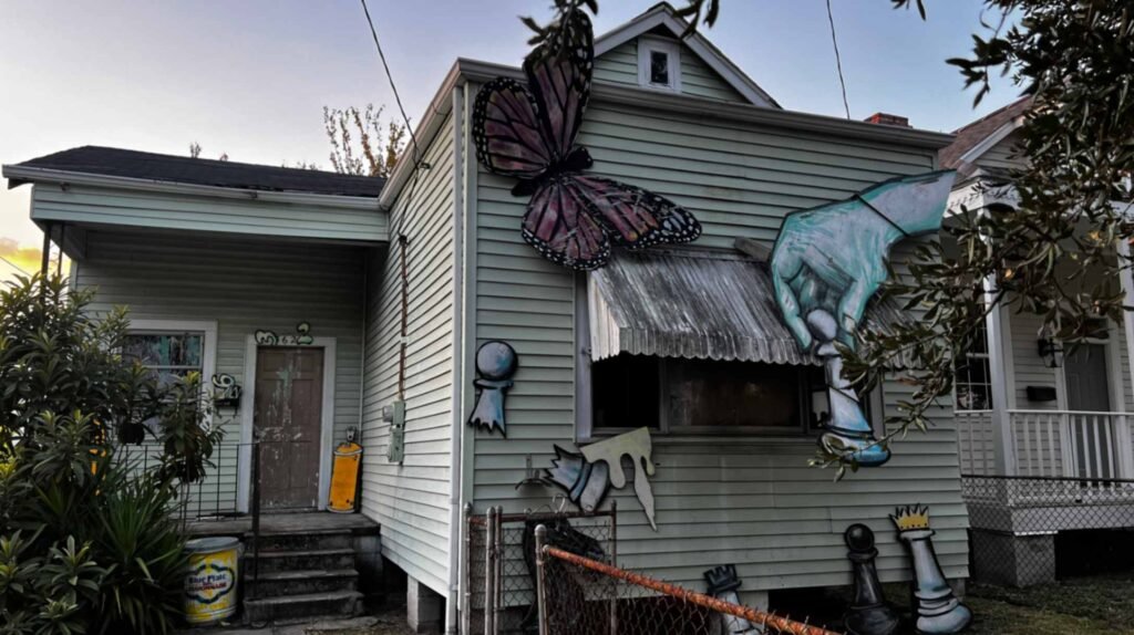 A light green residential house decorated with large, 2D pop-art cutouts including a giant butterfly, a hand holding a chess piece, and various chess pieces leaning against the foundation.