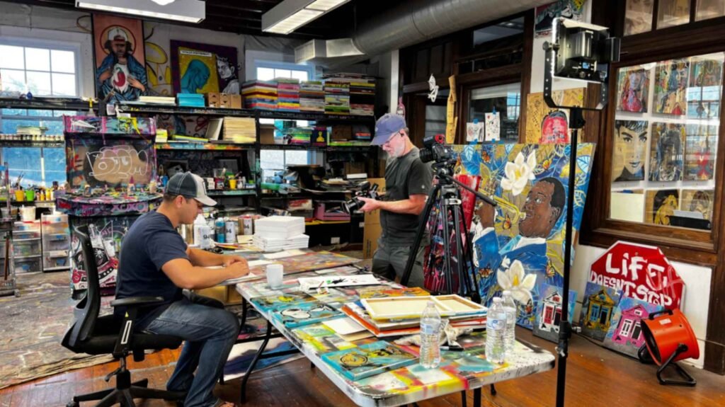 Joshua Wingerter sitting at a paint-splattered table in his studio, working on a canvas while a photographer captures the process. The room is filled with stacks of art and a large portrait of Louis Armstrong.