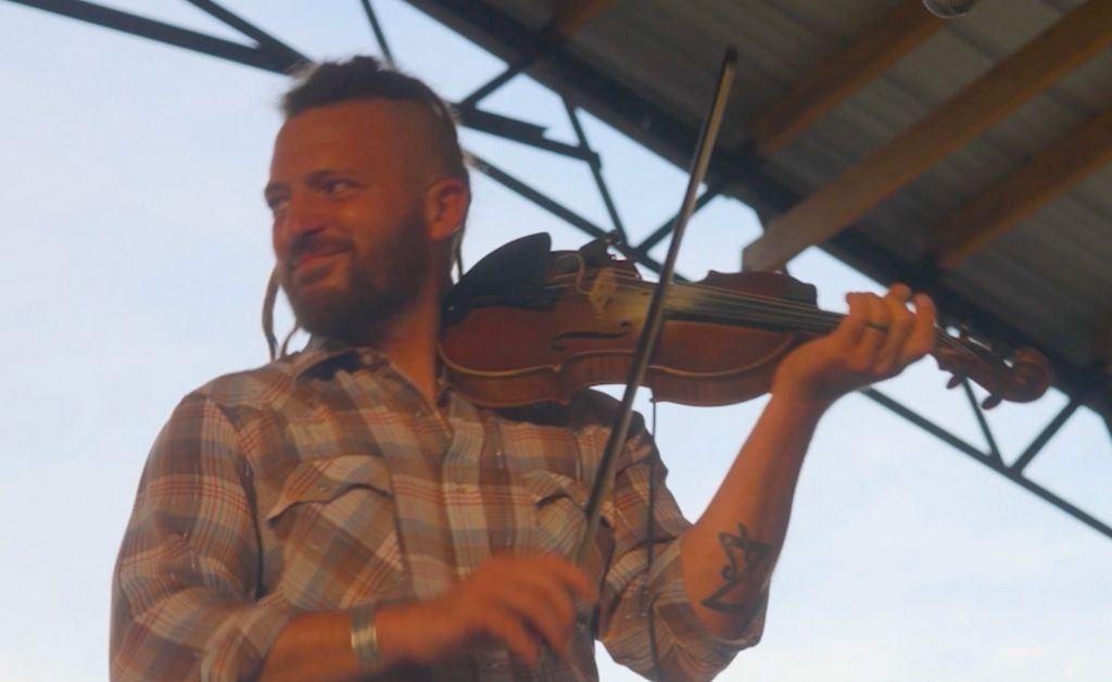 ourdan Thibodeaux smiling while playing the fiddle, showing his signature hairstyle and arm tattoo.