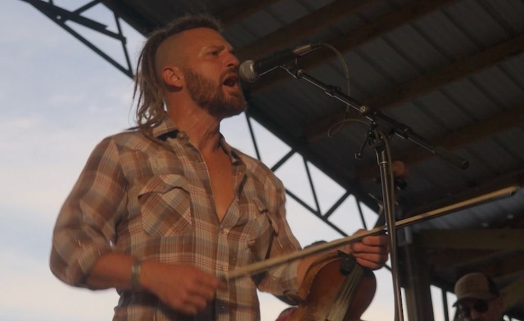 Jourdan Thibodeaux singing passionately into a microphone while holding his fiddle on stage.