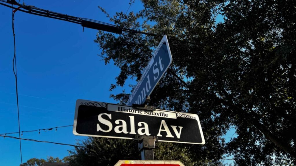 A black and white street sign for "Sala Av" in the Historic Salaville district of Westwego, Louisiana, set against a bright blue sky and lush green trees.