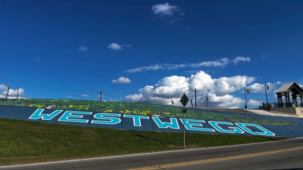 Large-scale "WESTWEGO" mural painted in bold blue and white block letters on a grassy levee slope under a bright blue sky with white clouds.
