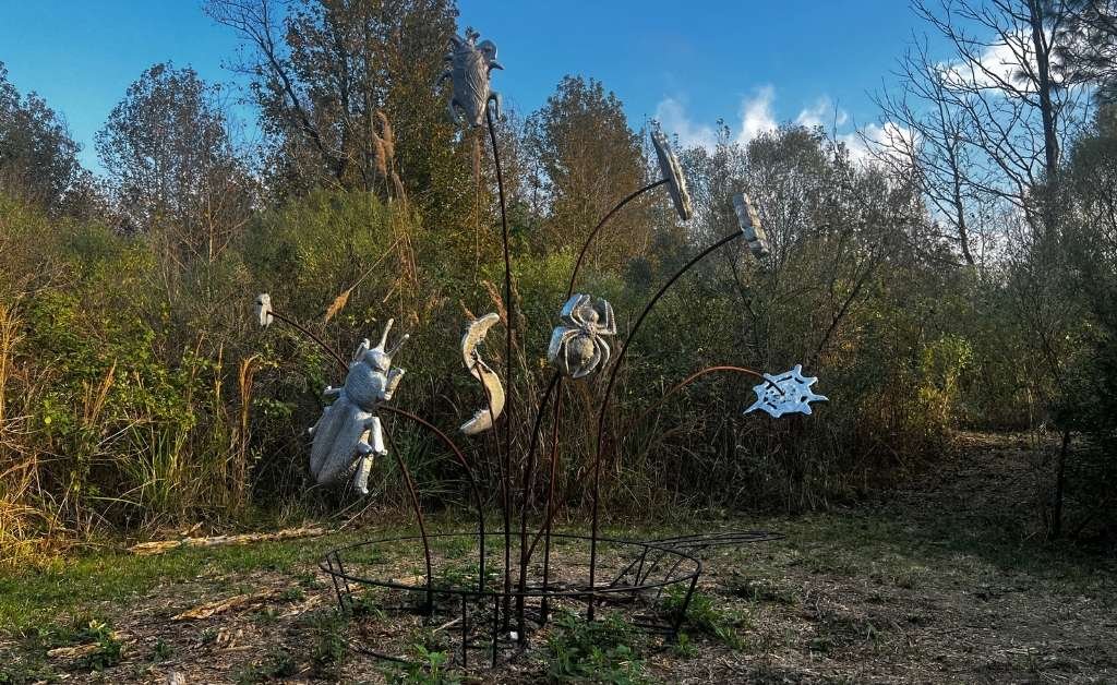 A large outdoor metal sculpture featuring various silver insect shapes, including a spider and a beetle, attached to the ends of long, thin metal rods that curve upward like a bouquet from a circular base at Atelier de la Nature.