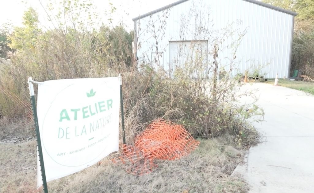 A white banner with the green "Atelier de la Nature" logo stands in front of a white metal building and natural brush.