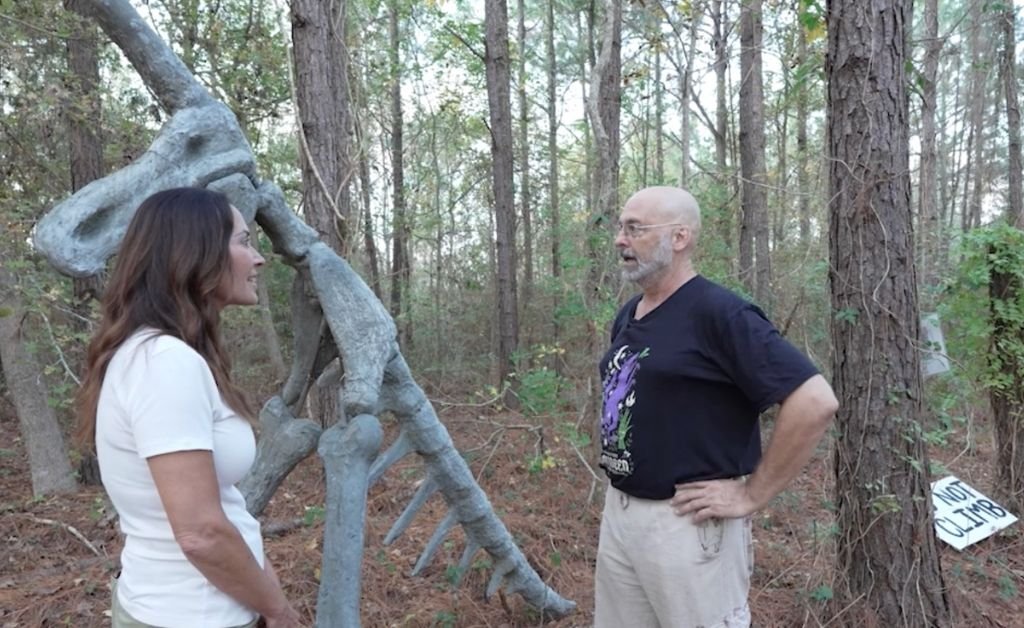 Karen LeBlanc stands beside a tall, gray, skeletal unicorn sculpture in a wooded area, speaking with a man in a black t-shirt at Atelier de la Nature.