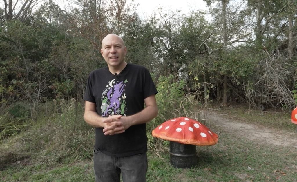Dr. Brandon Ballengée stands on a dirt path at Atelier de la Nature next to a large sculptural mushroom cap resting on a black barrel.