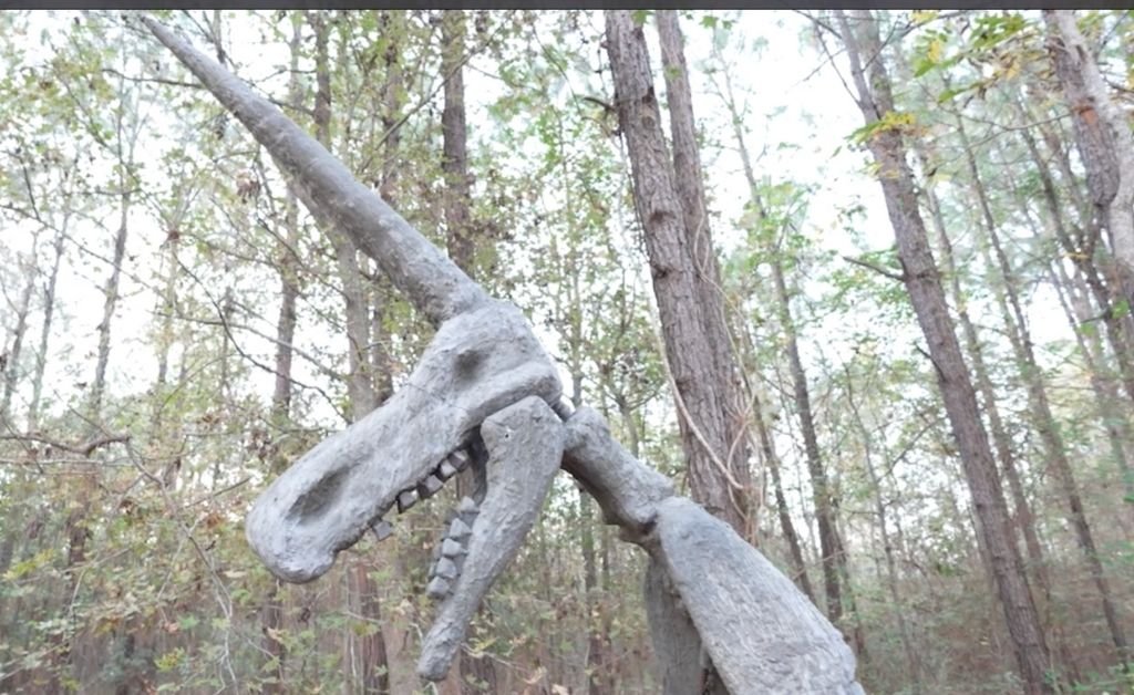 A large, gray, bone-like sculpture of the infamous Magdeburg Siberian unicorn standing among tall trees in a forest at Atelier de la Nature.