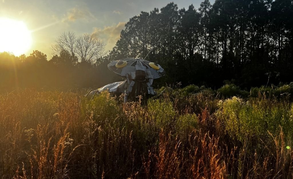 A large outdoor metal sculpture featuring various silver insect shapes, including a spider and a beetle, attached to the ends of long, thin metal rods that curve upward like a bouquet from a circular base at Atelier de la Nature.