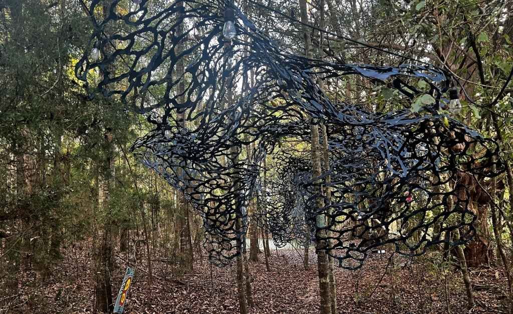 A large, black, net-like lattice structure is suspended between trees over a leaf-covered forest floor at Atelier de la Nature, with small light bulbs hanging nearby.