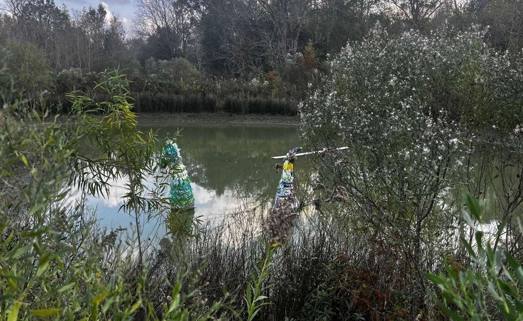 Two colorful sculptures made of recycled materials and glass bottles rise from a pond, framed by thick green vegetation and a backdrop of trees at Atelier de la Nature.