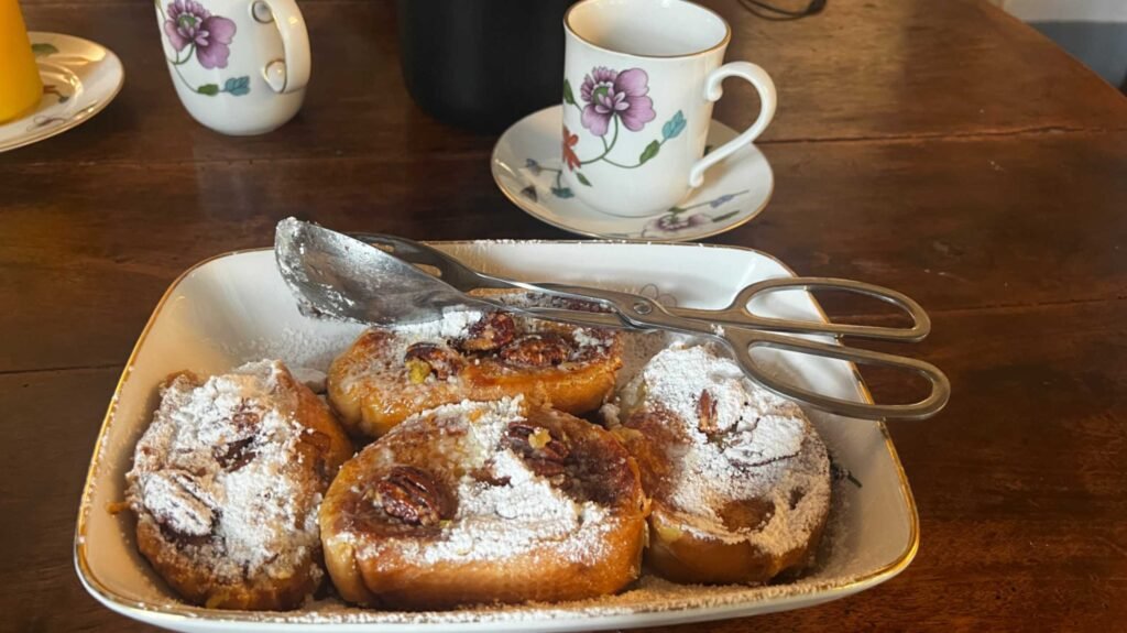 A white, gold-rimmed rectangular serving dish containing four thick slices of pain perdu (French toast) topped with whole pecans and a heavy dusting of powdered sugar. Metal serving tongs rest on the dish, and floral tea cups sit on a rustic wooden table in the background.