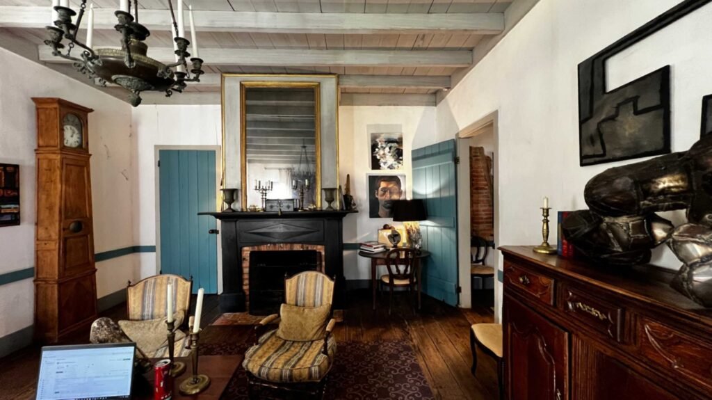 The interior of a historic French Creole cottage featuring a black fireplace mantel with a large gold-framed mirror, wooden beam ceilings, and wide-plank floors. The room is decorated with a mix of antique furniture, including a grandfather clock, and modern artwork, with Prussian blue shutters and doors.