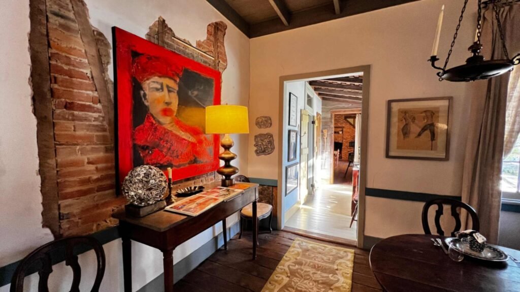 Text: An interior view of a cottage showing exposed brick-between-post wall construction and a large vibrant red painting. An antique wooden console table and a lit lamp sit on wide-plank wood floors.