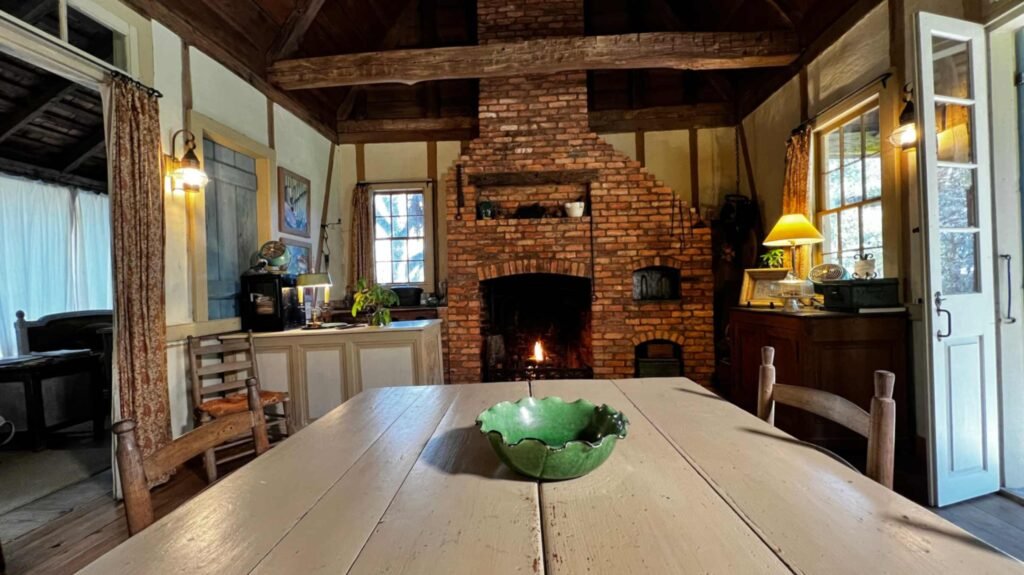 : A large, floor-to-ceiling rustic brick fireplace with a roaring fire, acting as the centerpiece of a dining room with exposed ceiling beams and a long wooden table.