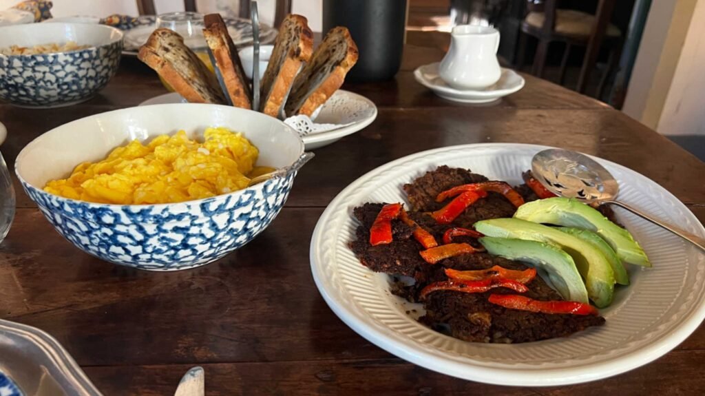 A breakfast spread on a dark wooden table featuring a blue and white patterned bowl of fluffy scrambled eggs, a plate of savory dark patties topped with roasted red peppers and fresh avocado slices, and a rack of toasted bread in the background.