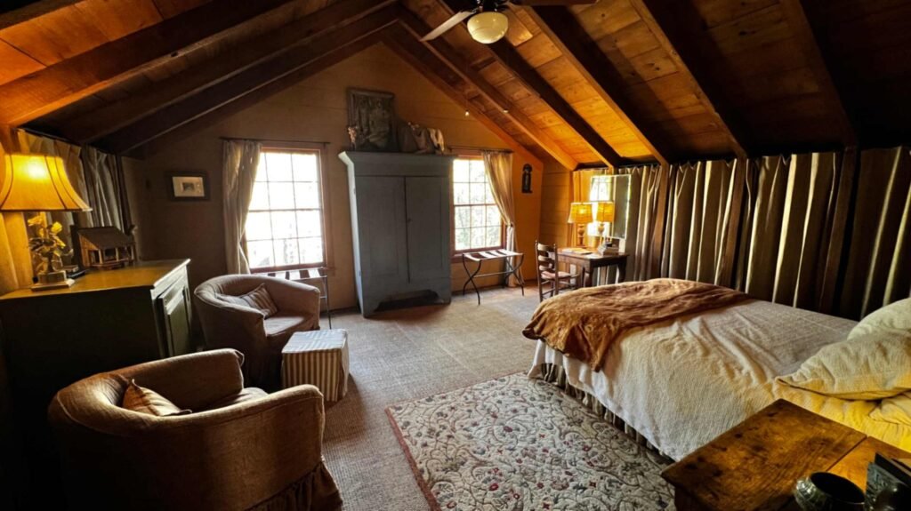 A spacious attic-level bedroom with a sloped wooden ceiling and exposed beams. The room features a large bed with a white and orange comforter, two armchairs, a large blue armoire, and patterned rugs.