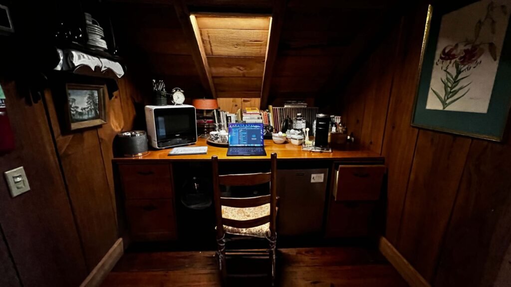 A cozy wooden desk setup under a sloped ceiling with a laptop, microwave, and coffee maker.