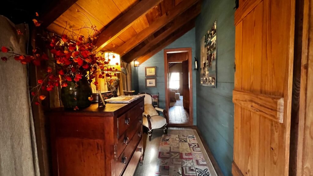 A narrow hallway with teal wooden walls, an antique wooden dresser, and a sloped ceiling leading to a guest room.