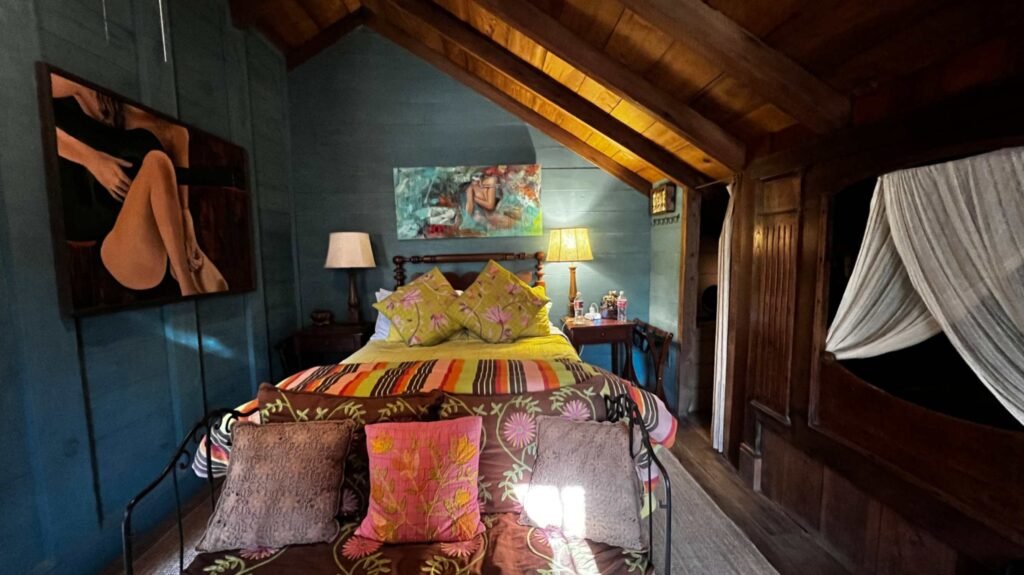 An antique-filled bedroom suite at Maison Madeleine featuring teal wooden walls, vibrant floral bedding, and modern art under a sloped wooden ceiling.