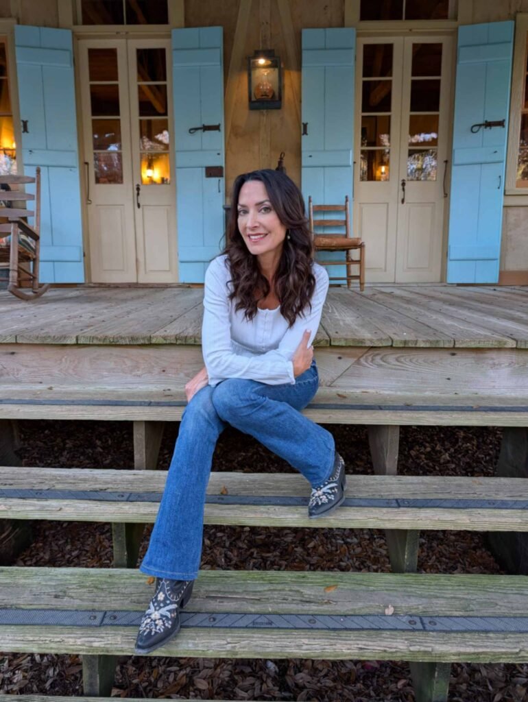 Host Karen sitting on the wooden steps of the wide front porch at Maison Madeleine, with light blue shutters and traditional French Creole architecture in the background.