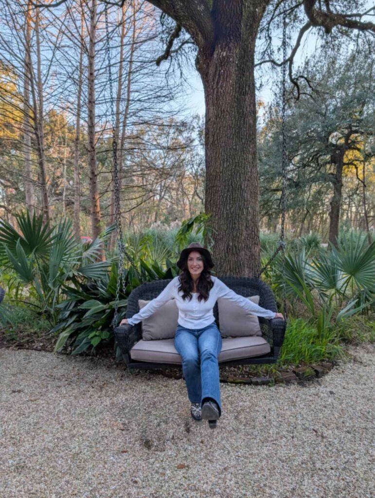 Karen wearing a hat and sitting on a black wicker porch swing hanging from a large tree in the gardens.