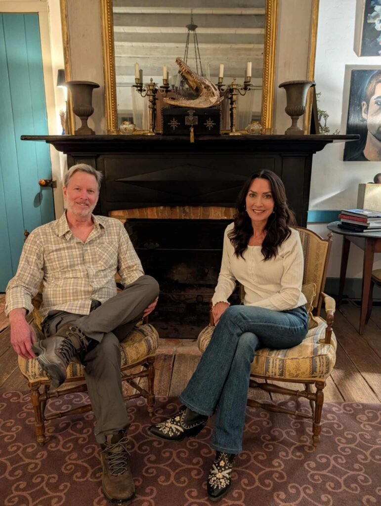 Ryan and Karen sitting in armchairs in front of a historic black fireplace with an alligator skull on the mantle.