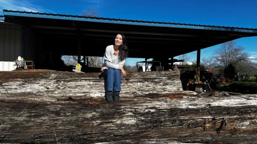 A woman sits centered on a single, enormous sinker cypress log that spans the width of the frame. Behind her is a rustic woodshop under a clear blue sky. The log’s bark is deeply grooved and grayed from age and water exposure.