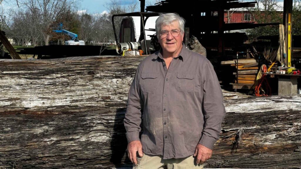 A high-angle view of a large, polished sinker cypress slab on a workbench. The wood features vibrant golden tones, swirling grain, and prominent dark mineral knots. Workshop tools and machinery are visible in the background.