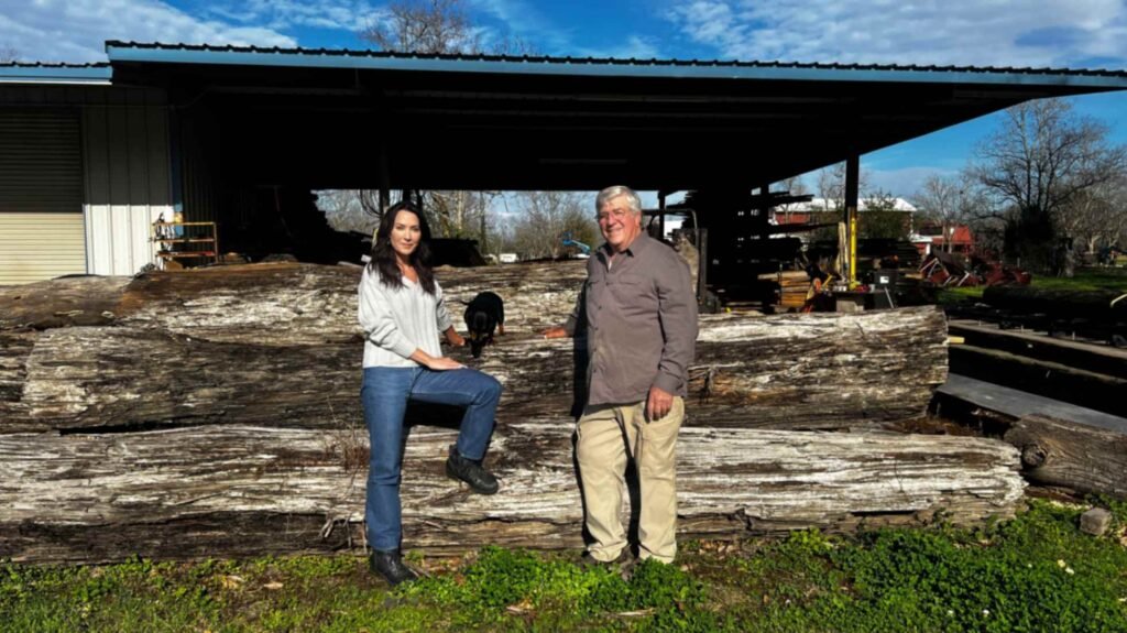 Louisiana craftsman Eric Couvillion and a female visitor standing in front of massive, long-reclaimed sinker cypress logs stacked outside a metal workshop. A small dog sits on one of the logs between them under a blue, slightly cloudy sky.