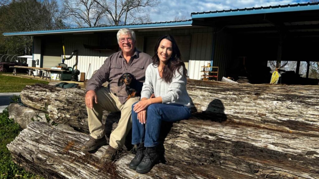 Louisiana craftsman Eric Couvillion, a woman, and a small dachshund sit atop a massive, weathered sinker cypress log outside a woodworking shop in the Atchafalaya Basin. The logs are thick, textured, and show the unique mineral staining of wood reclaimed from the swamp floor.