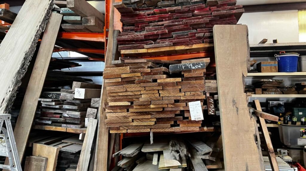 Neatly organized stacks of cut sinker cypress lumber in various shades of brown and red, stored on orange industrial shelving inside a workshop.