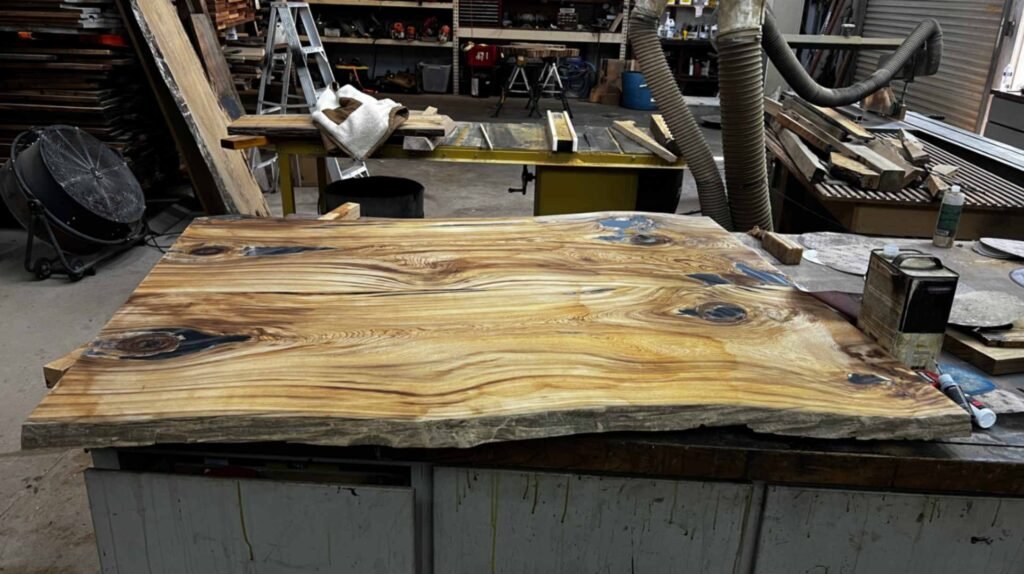 A high-angle, close-up view of a large, finished sinker cypress tabletop. The wood features vibrant gold and amber tones with dark, swirling knots and unique grey mineral deposits.
