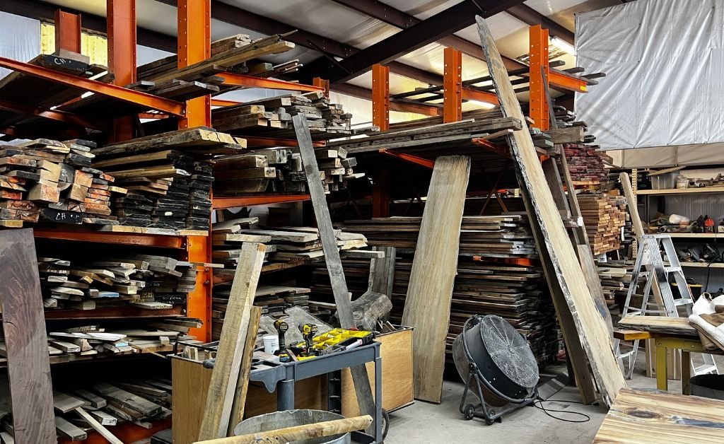 A wide interior shot of Eric Couvillion’s warehouse featuring floor-to-ceiling orange industrial cantilever racks stacked with hundreds of reclaimed wood planks and sinker cypress slabs. A large shop fan and various power tools on a cart are in the foreground.
