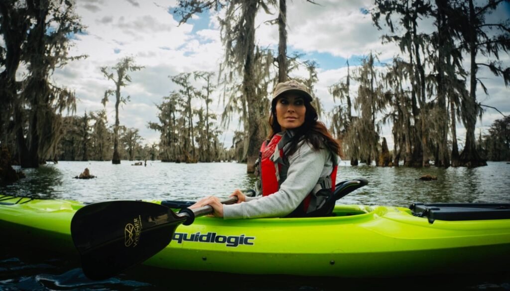 A woman in a grey sweatshirt and red life vest sits in a bright lime-green kayak, holding a black paddle. She is looking off-camera against a backdrop of Lake Martin's iconic cypress trees and hanging Spanish moss under a soft winter sky.